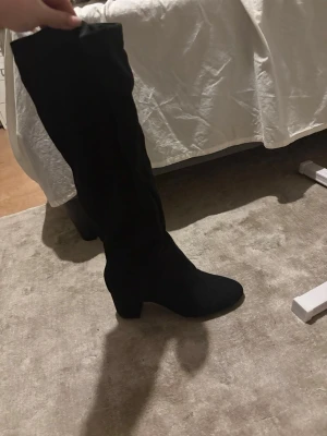 Svarta höga stövlar med klack - Säljer ett par svarta höga stövlar med låg klack. Skorna är i mjukt mockamaterial och har en stilren, klassisk look som passar till många outfits. Perfekt för dig som vill ha ett par snygga boots med lite höjd.