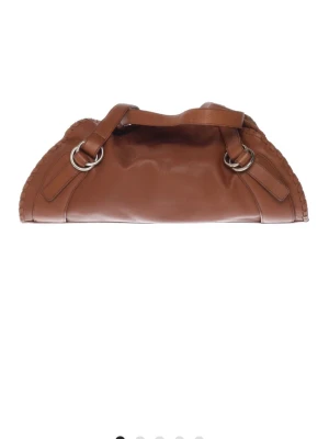 Brun skinnaxelväska Adax & Scotty - Säljer en brun axelväska från Adax & Scotty i mjukt skinn med snygga metallringar och dekorativa sömmar längs kanterna. Väskan har en rundad form och breda handtag, perfekt för dig som gillar stilrena detaljer.