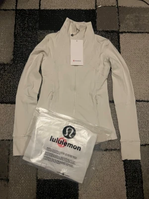 Ljusbeige zip-tröja från Lululemon - Supersnygg ljusbeige zip-tröja från Lululemon med hög krage och figurnära passform. Tröjan har dragkedja framtill, praktiska fickor med zip och coola sömdetaljer. Perfekt för träning eller chill. Materialet är mjukt och stretchigt syntet för maximal komfort.