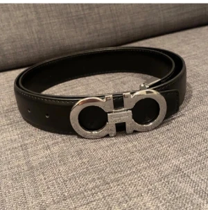 Ferragamo Gancini  - Säljer ett stilrent svart bälte från Ferragamo med ikoniskt silverfärgat spänne i metall. Bältet är tillverkat i läder och har en elegant design som lyfter vilken outfit som helst. Perfekt för dig som gillar exklusiva accessoarer.