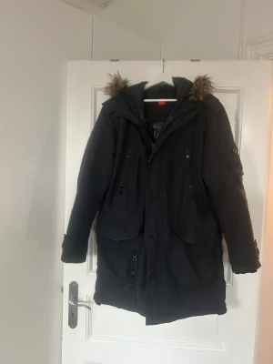 Bossjacka  - En varm och väldigt välvårdad Boss jacka, längre modell med huva. Jackan har 7 fickor på utsidan ( 3 st är lite mindre) och 3 innerfickor