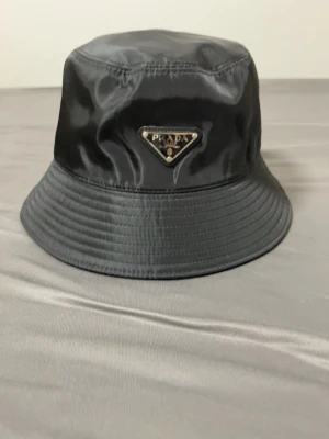 Svart bucket hat från Prada - Snygg svart bucket hat från Prada med ikonisk metalltriangel framtill. Hatten är tillverkad i polyester med bomullsfoder och har en stilren, glansig finish. Perfekt accessoar för att lyfta din look med lite lyxig streetstyle.helt ny kommer med lappar och en plastpåse till den tar byten bror på vad och fraktar genom plick 