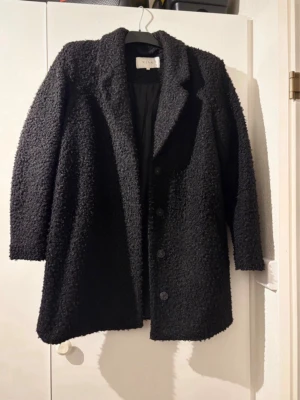 Svart teddyjacka från Vila - Mysig svart teddyjacka från Vila med knappar framtill och klassisk krage. Jackan har en grov, fluffig struktur och är långärmad. Perfekt för dig som gillar en chill och trendig look under kyliga dagar.