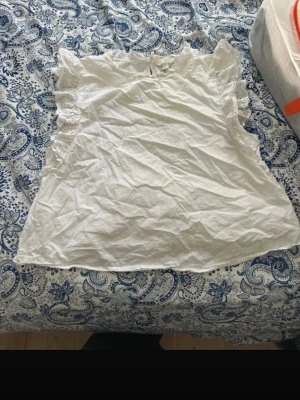 Vit blus med spetsvolang från H&M - Säljer en söt vit blus från H&M i storlek L. Blusen har spetsvolanger längs axlarna och är ärmlös. Tillverkad i bomull och har en rund halsringning med liten volangkant. Perfekt för sommarens alla tillfällen!