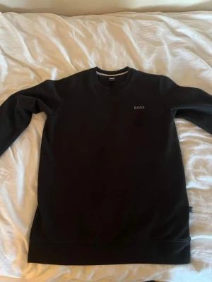 Svart sweatshirt från BOSS - Svart sweatshirt från BOSS med diskret logga på bröstet. Tröjan har rund halsringning och långa ärmar. Materialet känns mjukt och är troligtvis bomull eller polyester. Passformen är normal och stilen är enkel och clean.