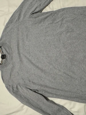 Grå stickad tröja från BOSS XXL - Snygg grå stickad tröja från BOSS i storlek L nu efter som att den har krympt i storlek, tillverkad av finaste italienska garnet. Tröjan har rund hals med ribbad kant och subtilt BOSS-logo broderat på bröstet. Perfekt för dig som gillar stilrena och klassiska plagg. Snabb affär 
