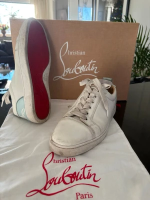 Christian Louboutin vita sneakers - Snygga vita sneakers från Christian Louboutin med klassisk röd sula och ljusblå detalj med logga på hälen. Skorna har rund tå, snörning och är tillverkade i skinn. Perfekt för dig som gillar exklusiva märken och stilren design. Skorna är 100% äkta och är köpte i Milano
