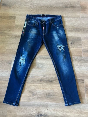 Mörkblå Dsquared2 jeans med slitningar - Snygga mörkblå jeans från Dsquared2 med coola slitningar och detaljer på benen. Modellen har klassisk femficksdesign, smal passform och tydliga kontrastsömmar. Materialet är jeans och byxorna har en modern, ungdomlig vibe.