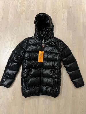 Svart pufferjacka från Save The Duck - helt ny med etiketter, originalkvitto medföljer från SkiStar, köpt till vår dotter som ångrade sig!Säljer en svart, glansig pufferjacka från Save The Duck med huva och två sidofickor med dragkedja. Jackan är quiltad och har en hel dragkedja framtill. Materialet är syntet och jackan är djurvänlig enligt märkningen på insidan.