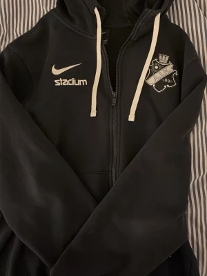 Svart AIK hoodie från Nike, stl S - Svart hoodie från Nike med AIK-emblem och Stadium-tryck på bröstet. Tröjan har vit snörning i huvan, dragkedja framtill och är långärmad. Perfekt för dig som gillar fotboll och vill ha en sportig stil.