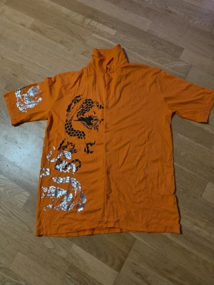 Orange skjorta med draktryck   - Cool orange skjorta med korta ärmar och knappar framtill. Har ett stort svart och silverfärgat draktryck på framsidan och ena ärmen. Skjortan har klassisk krage och är gjord i mjukt stretch  Perfekt för dig som vill sticka ut med färg och mönster.