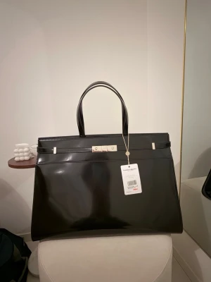 Svart handväska från Saint Laurent - Säljer en stor och stilren svart handväska från Saint Laurent Paris. Väskan är i glansigt skinn med silverfärgade metalldetaljer och har två rejäla handtag. Klassisk och elegant design med Saint Laurent-logga på låset framtill.Köpt från Vestiaire och den är authenticated. Sold out model. Original pris över 35000kr. 