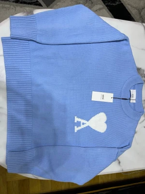 Ami Paris tröja - Snygg ljusblå stickad tröja från Ami Alexandre Mattiussi med ett vitt hjärtmönster och A-logga på bröstet. Tröjan har rund hals, ribbade muddar och är långärmad. Perfekt för dig som gillar stilrena detaljer och vill ha något som sticker ut.