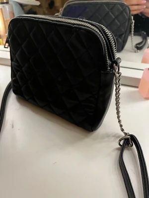 Svart quiltad axelväska med kedja - Säljer en svart quiltad axelväska med silverfärgad kedja och dubbla dragkedjor. Väskan har en stilren, rektangulär form och är tillverkad i skinnimitation. Perfekt för dig som gillar klassisk och trendig design med snygga detaljer.