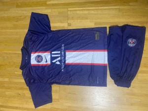 PSG Mbappé fotbollströja och shorts - Säljer ett mörkblått PSG matchställ med Mbappé 7 på ryggen. Tröjan har vita och röda detaljer, PSG-logga och Nike swoosh framtill samt sponsortryck. Shortsen är mörkblå med PSG-emblem. Perfekt för dig som älskar fotboll och Paris Saint-Germain.