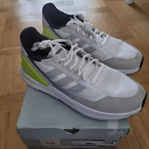 Adidas sneakers med vit och neon - Fräscha Adidas sneakers med vit mesh och gråa detaljer, samt inslag av neon-gult vid hälen. Skorna har klassiska tre ränder på sidan, svart innersula och snörning. Sulan är tjock och dämpad med svart och neon-gult undertill. Perfekt för dig som gillar sportig stil.