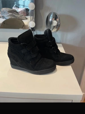 Nelly skor - Säljer ett par svarta sneakers med kilklack och tre breda kardborreband. Skorna är gjorda i mocka och har en platt sula framtill samt rund tå. 