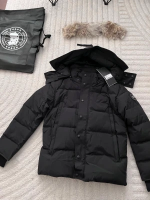 Svart pufferjacka från Canada Goose - Svart pufferjacka från Canada Goose med avtagbar huva och pälsdetalj. Jackan har quiltad design, dragkedja med logotyp och tryckknappar framtill. Märkets patch på ärmen ger en clean look. Perfekt för kalla dagar och riktigt snygg till streetwear.