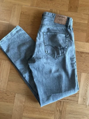 Ljusblå jeans från Jack & Jones - Säljer ett par ljusgrå jeans från Jack & Jones med klassisk femficksdesign och läderpatch bak i midjan. Jeansen har raka ben och är tillverkade i mjukt denimtyg som ger en avslappnad vibe. Perfekta till sneakers eller boots.