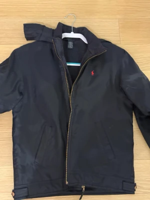 Svart vindjacka från Polo Ralph Lauren - Svart vindjacka från Polo Ralph Lauren med röd logga på bröstet. Jackan har huva, dragkedja framtill och två snedställda fickor. Materialet är nylon med polyesterfoder och fyllning. Perfekt för blåsiga dagar och enkel att matcha med din stil.