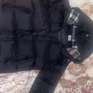 Svart pufferjacka från Burberry - Svart pufferjacka från Burberry med rutigt foder i svart, vitt och rött. Jackan har huva med spänne, dragkedja och knappar framtill samt praktiska fickor med dragkedja. Yttermaterialet är polyester och passformen är normal. Blir till väst, Mycket skön och fin. Varm och perfekt till vintern. Priset kan diskuteras. Digitalt kvitto finns. /Mvh