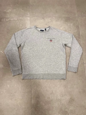 Grå sweatshirt från GANT - Snygg grå sweatshirt från GANT med broderad logga och emblem på bröstet. Tröjan har rund hals, ribbade muddar vid ärmslut och nederkant samt långärmad passform. Perfekt för chill eller skola, enkel att matcha med jeans eller joggers.