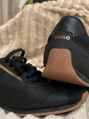 Svarta sneakers från Hugo med bruna detaljer - Säljer ett par svarta sneakers från Hugo med snygga bruna detaljer längs sidorna och på sulan. Skorna har rund tå, platt sula och klassisk snörning. Materialmix av skinn och syntet ger en cool look. Hugo-loggan syns både på hälen och innersulan.