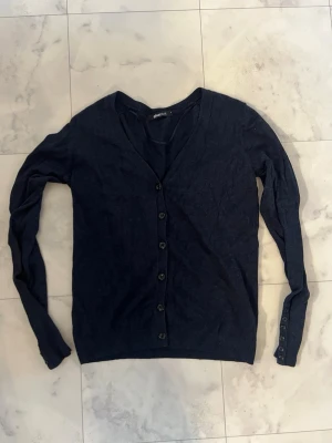 Mörkblå kofta från Gina Tricot - Snygg mörkblå kofta från Gina Tricot i storlek S. Koftan har v-ringad hals, långa ärmar och knappar hela vägen framtill samt vid ärmsluten. Perfekt att slänga över en t-shirt eller linne för en chill look.