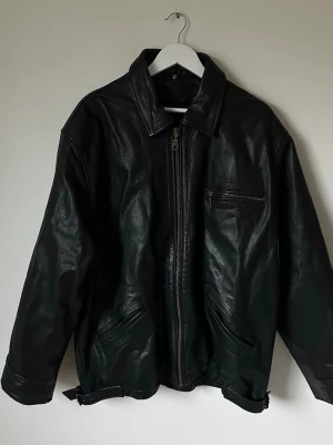 Svart skinnjacka med dragkedja - Svart skinnjacka med klassisk krage och grov dragkedja framtill. Jackan har tre fickor med dragkedja och justerbara spännen vid nederkanten. Insidan är quiltad för extra komfort och värme. Perfekt för dig som gillar en cool och tidlös stil. Jackan är i mycket bra skick men dragkedjan på bröstfickan trasig, se bild. 6Q2