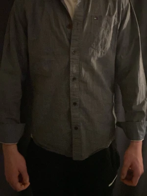 Grå skjorta från Tommy Hilfiger - Snygg grå skjorta från Tommy Hilfiger med klassisk krage och bröstficka med liten logga. Skjortan har långa ärmar, knappar framtill och en diskret detalj i nacken. Perfekt för dig som gillar stilrena och enkla plagg.