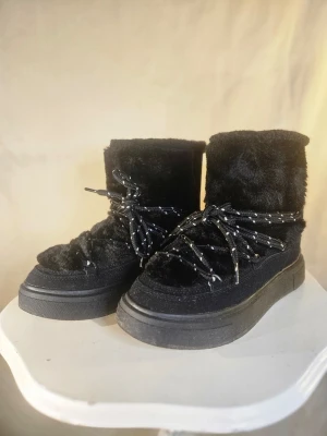 Svarta fluffiga moon boots med snörning - Säljer ett par svarta boot med fluffigt pälsmaterial och grov snörning med vita detaljer. Skorna har en platt sula och rund tå, perfekt för kalla dagar. De är höga och har en chunky look som sticker ut. Sulan är robust.