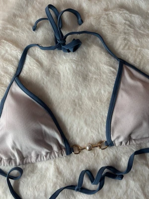 Blå och vit bikiniöverdel med pärldetalj - Bikiniöverdel i ljusblått och vitt med ribbad struktur. Har blå kanter och knytband både i nacken och runt ryggen. Mellan kuporna sitter en guldfärgad detalj med en pärla.