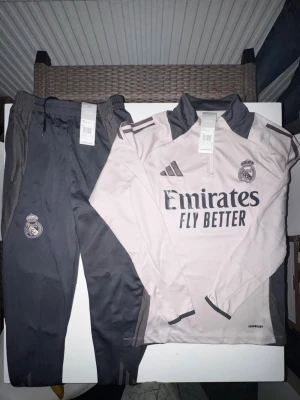 Adidas x Real Madrid fotbollströja med byxor - HELT NYA! Adidas Real Madrid träningsset i storlek M, med långärmad tröja i ljusbeige och grått samt matchande mörkgrå träningsbyxor. Tröjan har halv dragkedja, broderade loggor och Aeroready-material. Byxorna har snörning i midjan och Real Madrid-emblem.