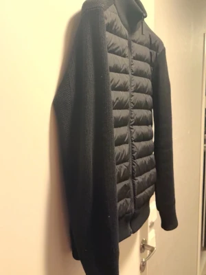 Svart hybridjacka från Canada Goose - Säljer en svart hybridjacka från Canada Goose med quiltad front och stickade ärmar och rygg. Jackan har dragkedja hela vägen, hög krage och den klassiska loggan på ärmen. Perfekt för dig som vill ha en stilren och funktionell jacka.