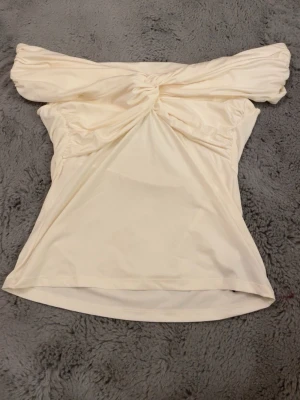 Offshoulder cremefärgad topp - Säljer en snygg offshoulder topp i cremevitt med knyt framtill. Toppen har en axelbandslös design och är gjord i ett mjukt syntetmaterial som sitter bekvämt mot kroppen. Perfekt för dig som gillar en feminin och trendig stil. Kan eventuellt tänka mig gå ner i pris
