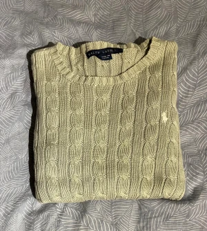 Beige stickad tröja från Ralph Lauren - Snygg beige stickad tröja från Ralph Lauren med klassiskt kabelstickat mönster och rund hals. Tröjan har långa ärmar och en liten broderad logga på bröstet. Perfekt för dig som gillar stilrena och tidlösa plagg.