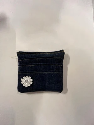 Mörkblå jeansplånbok med blomma - Säljer en liten plånbok i mörkblå denim med gul söm och dragkedja upptill. Framsidan har en vit broderad blomma som detalj. Perfekt för kort och småsaker, enkel och stilren design med en touch av personlighet.