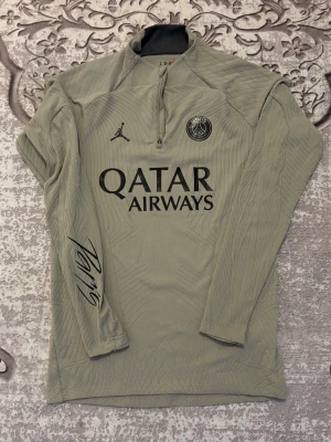 Jordan PSG half zip tröja slim fit - Snygg ljusgrå långärmad tröja från Jordan PSG med half zip och svart Paris-tryck på ärmen. Tröjan har broderad PSG-logga, Jordan-logga och Qatar Airways-tryck på bröstet. Slim fit och tillverkad i polyester med coolt mönster i tyget.