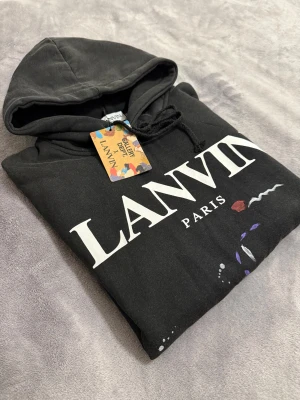 Lanvin tröja  - Svart hoodie från Lanvin x Gallery Dept med stor vit logga på bröstet och färgglada abstrakta detaljer framtill. Tröjan har huva med snörning och ribbade muddar vid ärmslut och nederkant. Perfekt för dig som gillar streetwear och unik design.