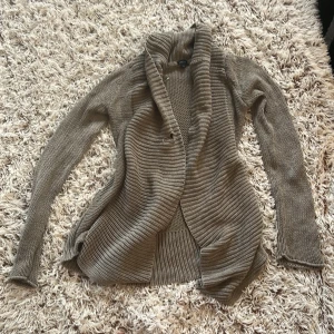 Beige stickad kofta från Lindex XS - Mysig beige stickad kofta från Lindex i storlek XS. Koftan har en öppen front med bred ribbad krage och långa ärmar. Perfekt för lager-på-lager och chill dagar. Tillverkad i bomull för en skön känsla. Perfekt till kalla nätter.  Tyvärr har den ett hål (se bild 2) Färgen är mörk beige 