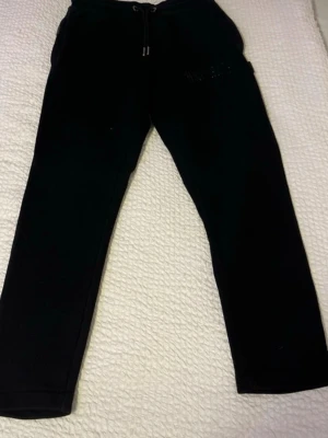 Svarta mjukisbyxor från Hoodrich - Svarta mjukisbyxor från Hoodrich med broderad logga på benet och snörning i midjan. Byxorna har en loose passform och är tillverkade i mjukt bomullsmaterial. Perfekta för chill eller träning, med bekväm midja och klassisk stil.