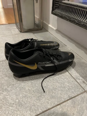 Nike Phantom GT fotbollsskor svart/guld - Säljer ett par Nike Phantom GT fotbollsskor i svart med snygga guldfärgade detaljer och silvriga mönster. Skorna har snörning och platt sula med dobbar för gräsplan och konstgräs. Materialet är syntet och designen är riktigt modern och sportig.
