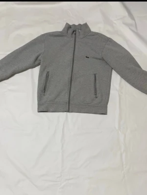 Grå fleecejacka från Lacoste - Säljer en grå fleecejacka från Lacoste i storlek M. Jackan har hel dragkedja framtill, två fickor med dragkedja och en liten Lacoste-logga på bröstet. Insidan är fodrad med mjuk teddyfleece som håller dig varm under kyliga dagar.
