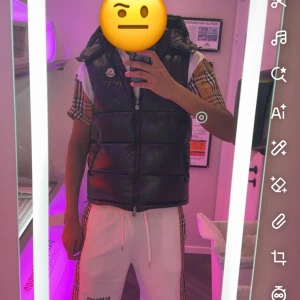 Svart dunväst från Moncler - Snygg svart dunväst från Moncler med glansig finish och hög krage. Västen har dragkedja framtill och ikonisk Moncler-logga på bröstet. Perfekt att slänga över en t-shirt eller hoodie för en sportig och trendig look. Storlek XL men passar M-L. NFC scan 