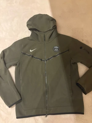 Olivgrön Nike PSG vindjacka med huva - Säljer en olivgrön Nike PSG vindjacka med huva och hel dragkedja. Jackan har PSG-logga och Nike Swoosh på bröstet, samt svarta detaljer längs kanterna. Tillverkad i bomull och polyester, perfekt för dig som gillar sportig stil.