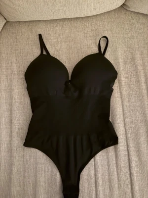 Svart body från H&M - Säljer en svart body från H&M i storlek M. Bodyn har vadderade kupor och smala axelband, vilket ger en snygg siluett. Materialet är mjukt och stretchigt, perfekt för att matcha med jeans eller kjol. Enkel och stilren design utan mönster. Skriv om ni har några frågor🥰