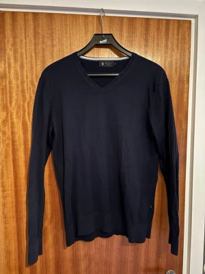 Mörkblå stickad tröja från Riley - Snygg mörkblå stickad tröja från Riley med v-ringad hals och långa ärmar. Perfekt för dig som gillar klassisk stil med enkel design. Tröjan är tillverkad i mjukt material och har ribbade muddar vid ärmslut och nederkant. Passar dig som vill ha en clean look. Tröjan är i mycket bra skick fast använd ett par fåtal gånger. Priset går att diskutera