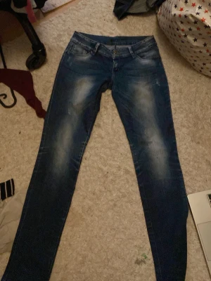 För lowriseBlå  jeans med broderad ficka - Snygga blå lowrise jeans med slitningar och coolt broderi med nitar på bakfickan. Jeansen har klassisk femficksdesign, knapp och dragkedja fram samt bälteshällor. Materialet är jeans och passformen är tight, perfekt för dig som gillar en smal siluett.