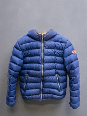 Blå pufferjacka från Colmar - Blå pufferjacka från Colmar med huva och två fickor med dragkedja. Jackan har ett quiltat mönster och Colmar-logga på ärmen. Materialet är polyester och jackan är perfekt för kyliga dagar. Jackan har 2 hål på ärmen och på ryggen men som har blivit lagade med lagnings lappar som ni ser på bild 2 och 7. Jackan har vanlig passform och sitter otroligt bra runt midjan och kropp