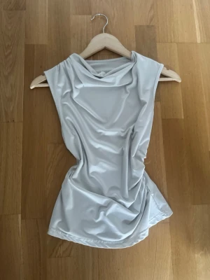Vit omlott ärmlös topp - Säljer en vit ärmlös topp med snygg omlott-drapering framtill. Toppen har en mjuk och glansig yta som ger en cool vibe och passar perfekt till jeans eller kjol. Materialet känns lätt och följsamt mot kroppen.